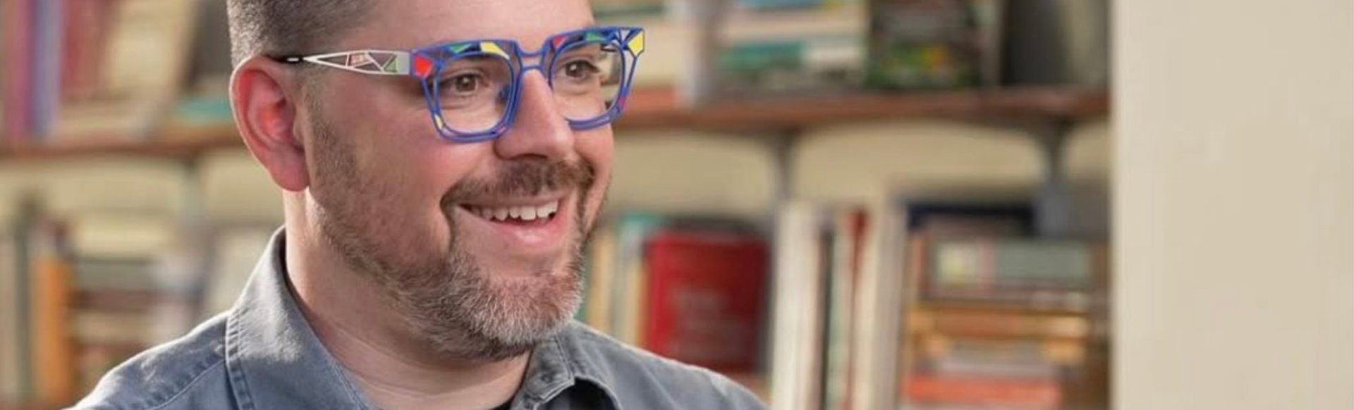 Danny Caine wearing colorful glasses, smiling with book lined shelves in the background.